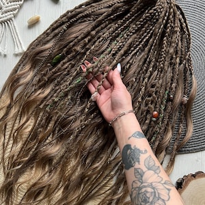 Peut inclure: Un ensemble d'extensions de cheveux synthétiques marron composé d'un mélange de dreadlocks et de mèches ondulées. Les cheveux sont ornés de petites perles décoratives et de fils verts, le tout attaché au sommet par un élastique neutre.