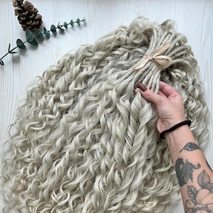 May include: A bundle of light gray, curly synthetic dreadlocks. The dreadlocks are tied together with a beige ribbon. The image shows the dreadlocks in a wavy pattern. The dreadlocks are a light gray color. The image is taken from a top-down perspective.