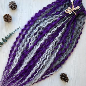 May include: A bundle of wavy synthetic dreadlocks in shades of purple, gray, and white. The dreadlocks are tied together with a beige ribbon. The image also includes pine cones and greenery.