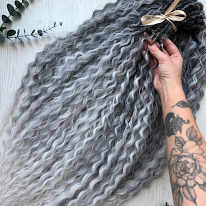Op de afbeelding: Grijze en witte golvende haarextensions, vastgebonden met een gouden lint. De extensions hebben een gradiënteffect, van donkergrijs naar wit. Een hand houdt een deel van de extensions vast, met een tatoeage op de arm. De achtergrond is een licht houten oppervlak.