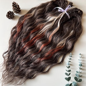 May include: A bundle of wavy, dark brown hair extensions with streaks of white, orange, and light brown. The extensions are tied together with a lavender ribbon. Three pinecones and sprigs of greenery are arranged around the hair.