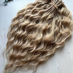 May include: A set of blonde synthetic hair extensions featuring wavy, loose curls. The hair strands are gathered at the top and secured with a small, light-colored ribbon tie, creating a cascading, voluminous appearance with varied golden tones.