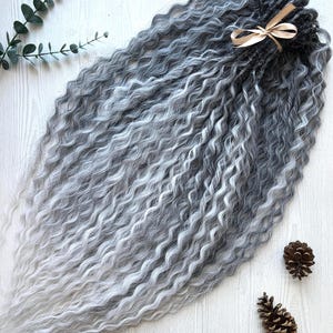 May include: A bundle of long, wavy synthetic hair extensions featuring an ombre color gradient that transitions from dark charcoal gray at the top to a light silver-white at the ends, secured at the base with a light beige ribbon bow.