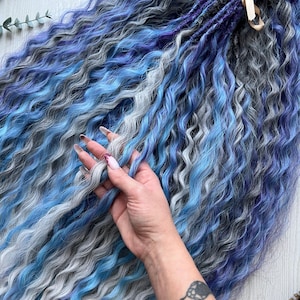 Könnte beinhalten: Ein Bündel gewellter, synthetischer Haarverlängerungen in Blau-, Grau- und Lilatönen. Das Haar ist mit einem goldenen Band gebunden. Die Extensions sind so konzipiert, dass sie dem natürlichen Haar Länge und Volumen verleihen.