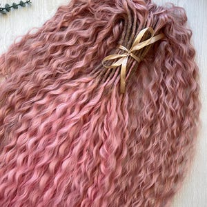 Puede incluir: Un conjunto de rastas sintéticas rosas con una textura ondulada. Las rastas están atadas con una cinta marrón claro. El color de las rastas varía de rosa claro a un tono rosa más oscuro.