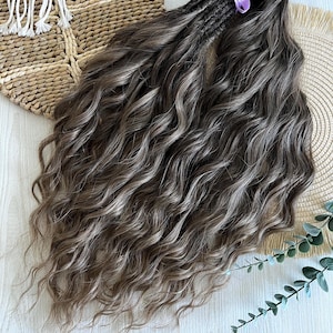May include: A set of long, wavy hair extensions in a dark brown and ash blonde highlighted color blend. The hair is gathered at the top and secured with a light purple ribbon, featuring a voluminous, textured appearance with loose curls.