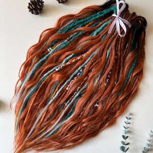 May include: A collection of wavy dreadlocks in copper, teal, and white, secured with a white ribbon. The dreads have decorative wraps and accents. Two pine cones are placed near the dreadlocks on a white surface.