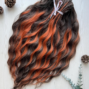 May include: Dark brown and orange wavy hair extensions with a white ribbon. The extensions have a mix of dark brown and vibrant orange strands, creating a striking contrast. The hair is styled in loose waves, and the extensions are tied together with a white ribbon.