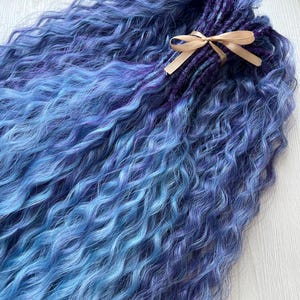 Puede incluir: Un conjunto de rastas sintéticas azules y moradas con textura ondulada, atadas con una cinta beige. El degradado de color va del azul claro al morado oscuro. Un accesorio para el cabello.