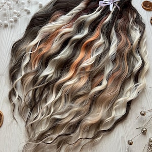 May include: A set of wavy hair extensions featuring a multi-tonal blend of dark brown, light brown, cream, and copper strands. The hair is gathered at the top and secured with a light purple ribbon, creating a cascading, textured appearance.