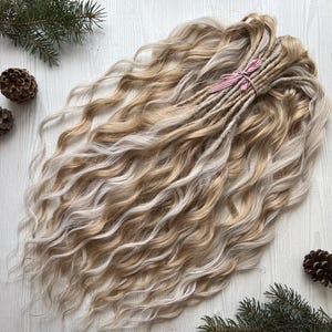 Peut inclure: Dreadlocks ondulées blondes et marron clair, attachées avec un ruban rayé rose et blanc. Les dreadlocks sont regroupées, avec des extrémités lâches et fluides. Les extensions de cheveux présentent un mélange de tons clairs et foncés.