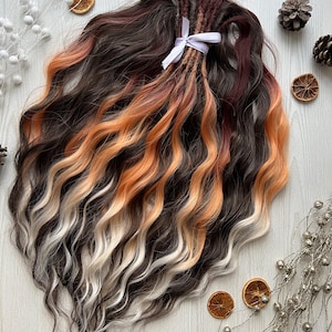 May include: A collection of wavy hair extensions in dark brown, auburn, orange, and white. The extensions are secured with a white ribbon. The hair is styled with loose waves, creating a natural and textured appearance.