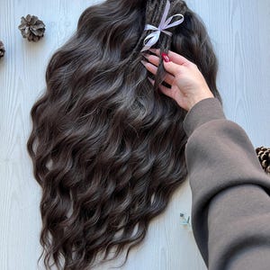 Op de afbeelding: Donkerbruine, golvende hairextensions met grijze highlights, vastgebonden met een lichtpaars lint. De extensions zijn ontworpen om er natuurlijk en volumineus uit te zien. De afbeelding toont een close-up van het haar, waarbij de textuur en kleurvariaties worden benadrukt.
