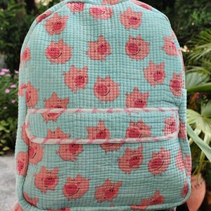 May include: A light blue quilted backpack featuring a repeating pattern of small pink cartoon pigs. The bag includes a front zippered pocket with a white and pink striped trim, a top handle, and a textured fabric surface throughout the exterior.