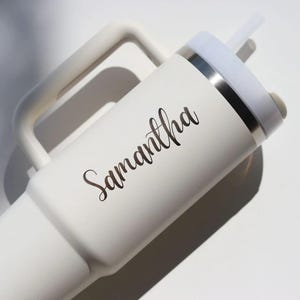 May include: White insulated tumbler with a handle and a straw. The tumbler has a white lid with a silver band and the name "Samantha" in a cursive font. The tumbler is designed to keep drinks cold or hot.