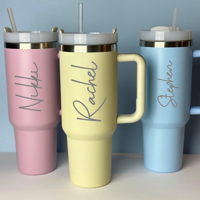 May include: Three insulated tumblers in pastel colors: pink, yellow, and blue. Each tumbler has a handle, a lid with a straw, and a name in a cursive font. The names are Nikki, Rachel, and Stephen.