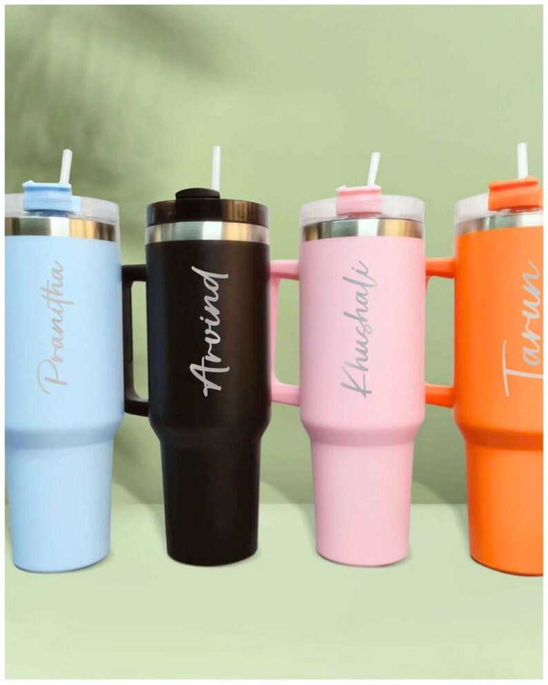 May include: Four insulated tumblers in pastel colors: light blue, black, pink, and orange. Each tumbler has a handle, a lid with a straw, and a personalized name in a cursive font. The tumblers are designed to keep drinks cold.