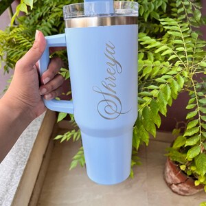 May include: A light blue insulated tumbler with a handle and a clear lid. The tumbler has the name "Shreya" engraved in a cursive font. The tumbler is shown against a background of green foliage.