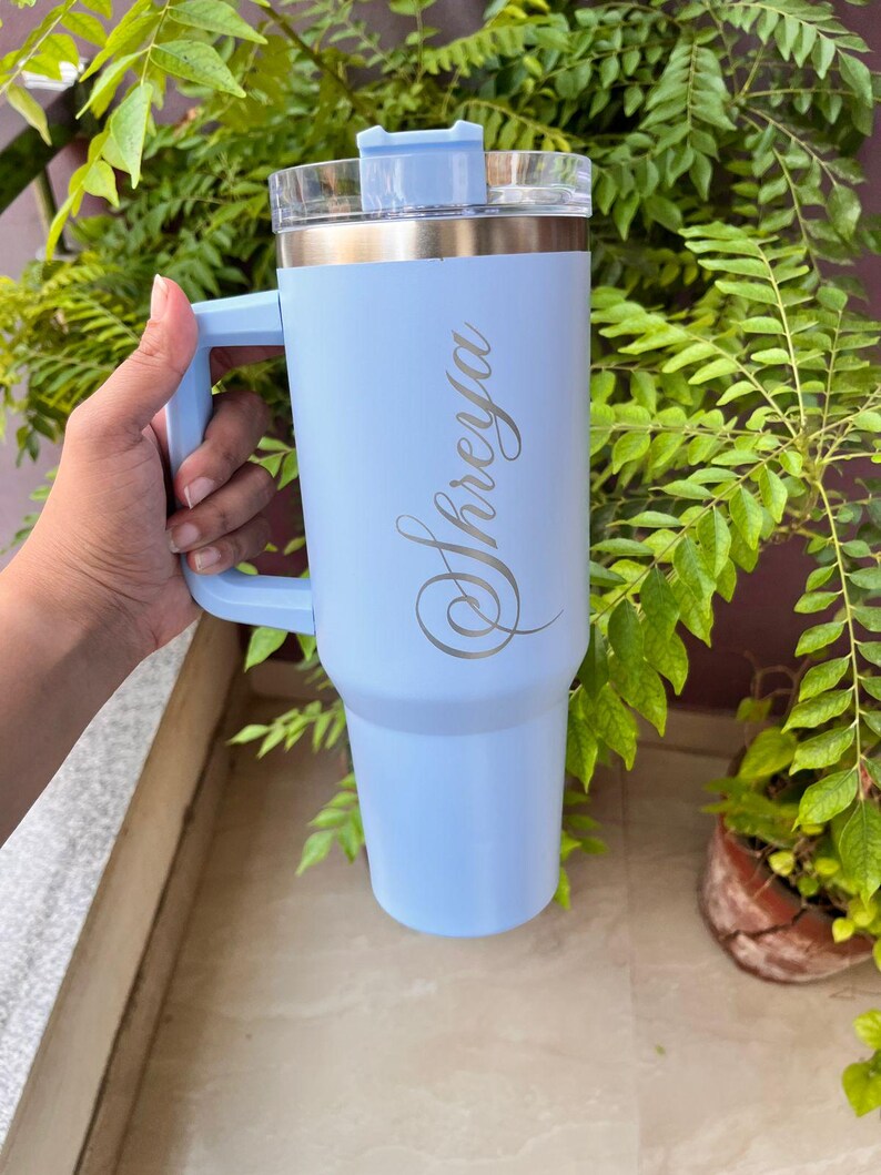 May include: A light blue insulated tumbler with a handle and a clear lid. The tumbler has the name "Shreya" engraved in a cursive font. The tumbler is shown against a background of green foliage.