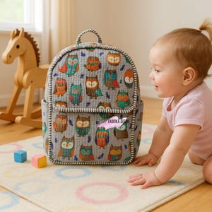 May include: A grey backpack featuring a pattern of colourful owls. The bag has a top handle and a front pocket. The edges are trimmed with a white and green striped fabric. The text "EXOTIKA ATTIRE" is visible on the front pocket.