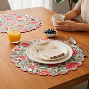 Op de afbeelding: Een tafelsetting met bloemen placemats, borden en een gevouwen servet. De placemats hebben een lichtblauwe achtergrond met rode bloempatronen en geschulpte randen. Een glas sinaasappelsap en een kom blauwe bessen zijn ook aanwezig.