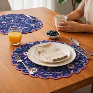 Può includere: Due tovagliette rotonde con sfondo blu scuro e un vivace motivo floreale nei toni del rosa, bianco e verde, rifinite con un bordo smerlato rosa chiaro. Ogni tovaglietta è apparecchiata con un piatto bianco, un tovagliolo beige e posate.