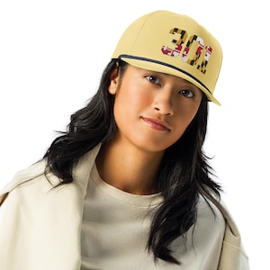 May include: A pale yellow cap featuring the number "301" in a Maryland flag pattern. The cap has a dark blue rope detail along the brim and a flat peak. The cap is worn by a person with dark hair.