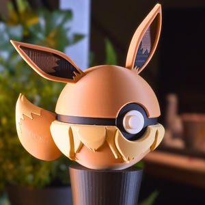 Könnte beinhalten: Eine 3D-gedruckte Eevee-Figur mit rundem, hellbraunem Körper, schwarzen Akzenten, großen Ohren und einem flauschigen Schwanz. Auf einem schwarzen zylindrischen Sockel montiert. Inspiriert von der Pokémon-Franchise.