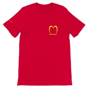 May include: A red t-shirt featuring a yellow logo resembling a stylized 'M' with the text "McDowell's" below it. The phrase "Say Mick!" is printed on the left sleeve. The shirt is a solid color and has a crew neck.