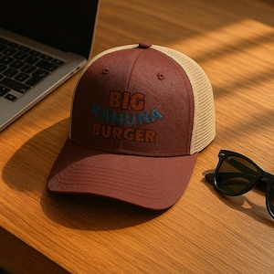 Könnte beinhalten: Eine bordeauxfarbene Trucker-Cap mit beigefarbenem Mesh-Rücken. Auf der Kappe steht der Text "BIG KAHUNA BURGER" in blauen und orangefarbenen Buchstaben. Eine schwarze Sonnenbrille liegt neben der Kappe auf einer Holzoberfläche.