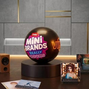 May include: A black spherical Zuru Mini Brands Really Works container sits on a circular black base. To the left is a small wooden speaker. To the right are two square album covers labeled Now Playing. Two additional album covers lie flat.
