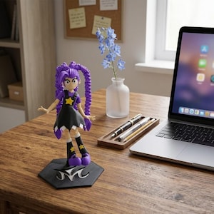 Op de afbeelding: Een anime-figuur met paars haar die op een zwarte zeshoekige voet staat. Het figuur draagt een zwarte jurk met een gele ster, paarse laarzen met gele sterren en heeft lang gevlochten haar. Het figuurtje staat op een houten bureau.