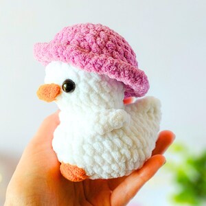 May include: A handmade white crochet duck wearing a pink hat. The duck has orange feet and beak, and black eyes. The plush toy is held in a person's hand.