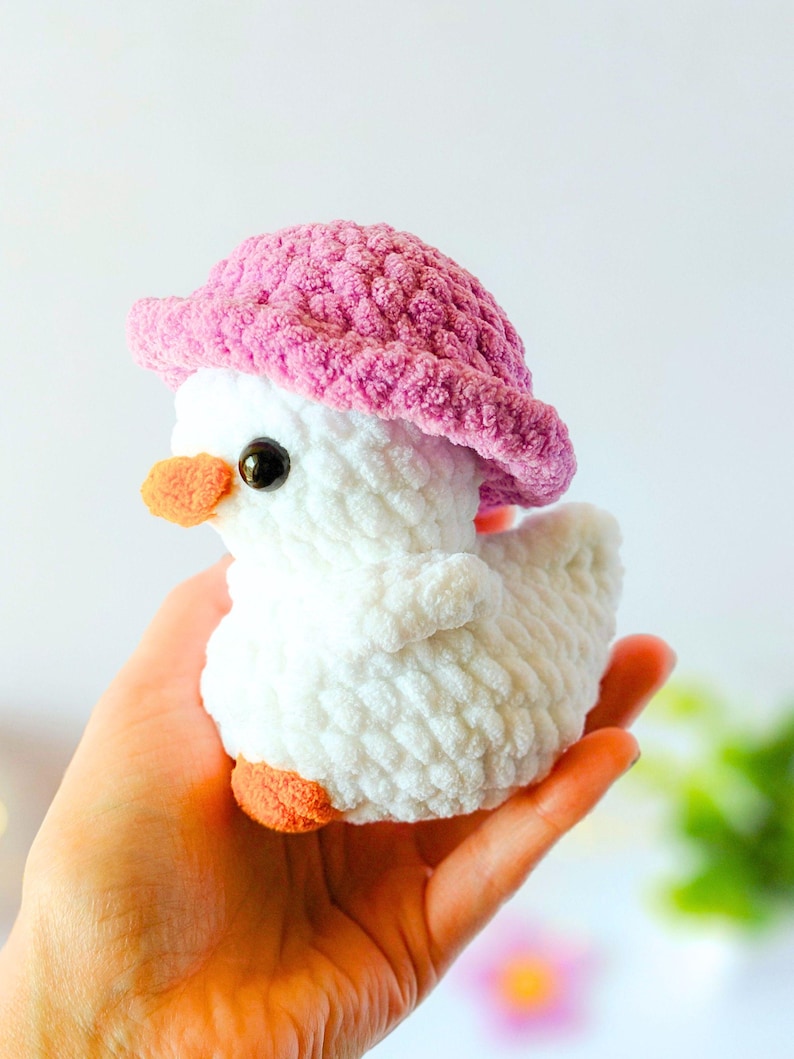 May include: A handmade white crochet duck wearing a pink hat. The duck has orange feet and beak, and black eyes. The plush toy is held in a person's hand.