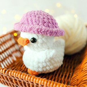 May include: A handmade white crochet duckling wearing a pink hat, with orange beak and feet, and black eyes. The duckling sits in a brown wicker basket, with a ball of white yarn in the background. A charming, whimsical decorative item.
