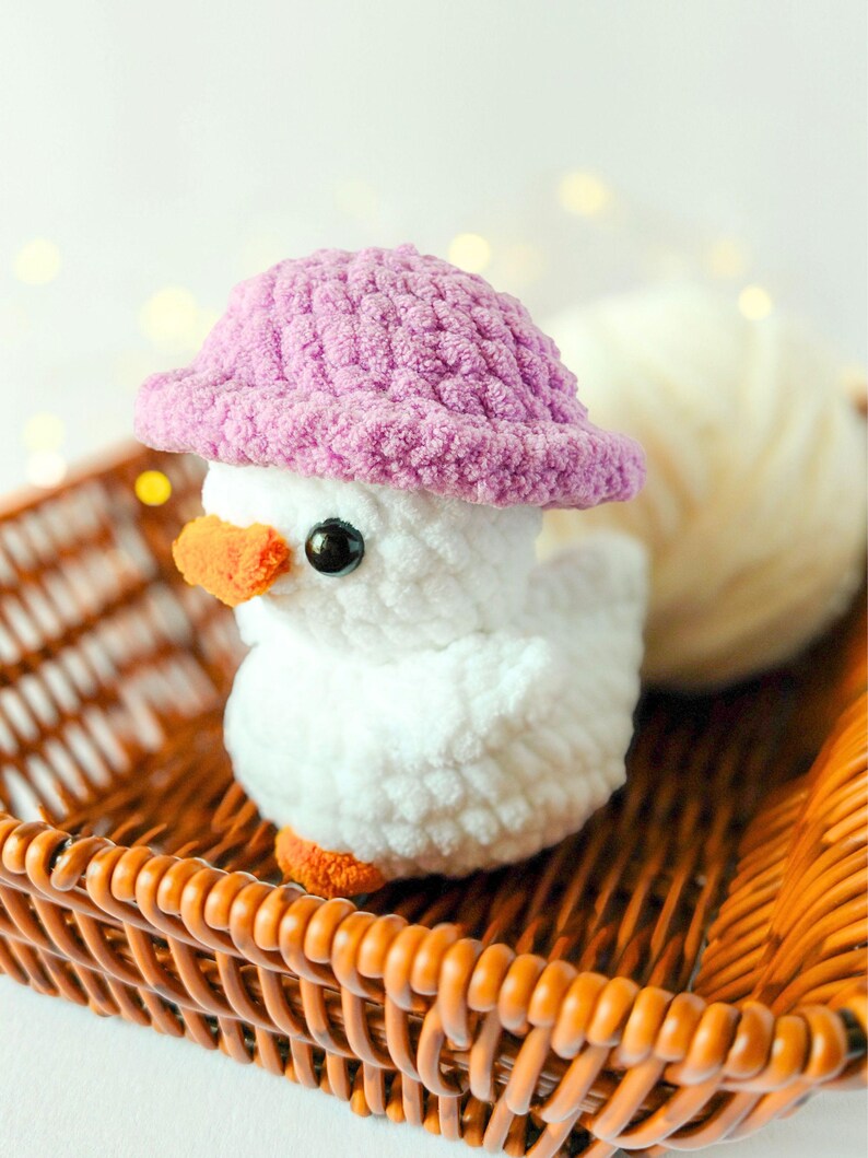 May include: A handmade white crochet duckling wearing a pink hat, with orange beak and feet, and black eyes. The duckling sits in a brown wicker basket, with a ball of white yarn in the background. A charming, whimsical decorative item.