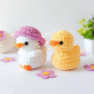 May include: Two crocheted ducklings, one white with a pink hat and the other yellow, sit on a white surface. Both have orange beaks and black eyes. Pink and yellow flower-shaped decorations are scattered around them.