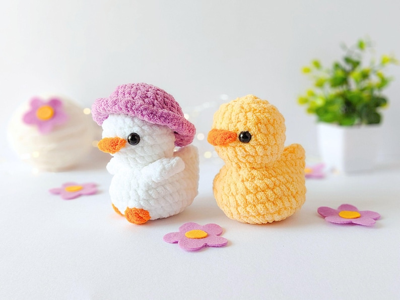 May include: Two crocheted ducklings, one white with a pink hat and the other yellow, sit on a white surface. Both have orange beaks and black eyes. Pink and yellow flower-shaped decorations are scattered around them.