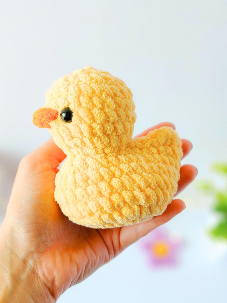 May include: A handmade, yellow crochet duck with a textured surface. The duck has a small orange beak and a black eye. The plush toy is a soft, cuddly item.