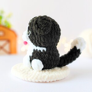 May include: A small, handmade crochet cat figurine featuring black and white yarn, a long tail with a white tip, and a small pink tongue. The cat is sitting on a circular, cream-colored crocheted base, showcasing detailed yarn texture throughout.