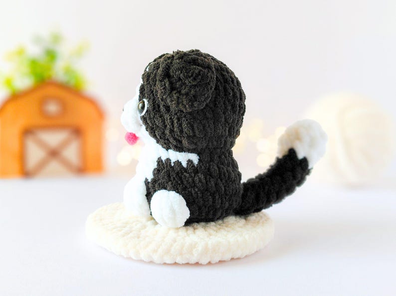 May include: A small, handmade crochet cat figurine featuring black and white yarn, a long tail with a white tip, and a small pink tongue. The cat is sitting on a circular, cream-colored crocheted base, showcasing detailed yarn texture throughout.