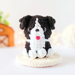 May include: A small, handmade crochet dog figurine featuring black and white yarn. The plush toy has a white face stripe, black ears, dark plastic eyes, and a small pink nose. It sits on a round, cream-colored crocheted base with a soft texture.