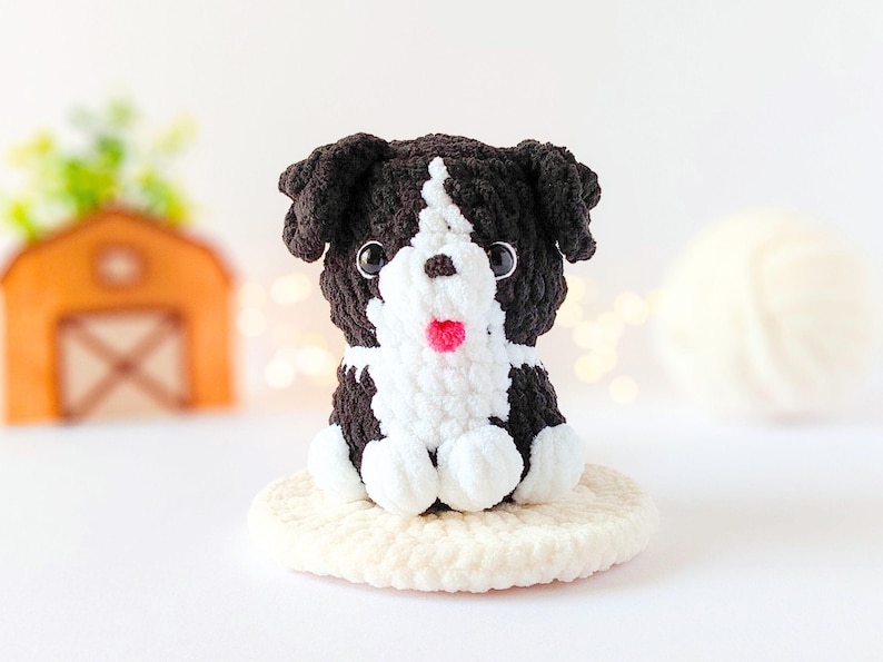 May include: A small, handmade crochet dog figurine featuring black and white yarn. The plush toy has a white face stripe, black ears, dark plastic eyes, and a small pink nose. It sits on a round, cream-colored crocheted base with a soft texture.