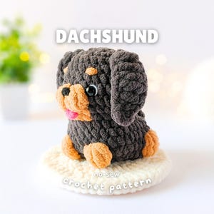 May include: A crocheted dachshund plush toy made of dark brown and tan yarn with black plastic eyes and a pink tongue. The text DACHSHUND and no-sew crochet pattern are visible above and below the plush, which sits on a round white base.