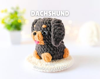 May include: A crocheted dachshund plush toy made of dark brown and tan yarn with black plastic eyes and a pink tongue. The text DACHSHUND and no-sew crochet pattern are visible above and below the plush, which sits on a round white base.