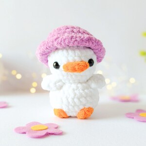 May include: A handmade white crochet duckling wearing a pink hat with an orange beak and feet. The duckling is surrounded by pink felt flowers with yellow centers. The background is white with soft bokeh lights.