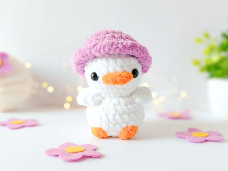 May include: A handmade white crochet duckling wearing a pink hat with an orange beak and feet. The duckling is surrounded by pink felt flowers with yellow centers. The background is white with soft bokeh lights.