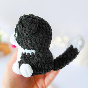 May include: A small, handmade crochet cat plushie featuring a black and white color scheme. The cat has a textured, fuzzy yarn body, a long tail with a white tip, small white paws, and a tiny pink tongue visible on its side profile.