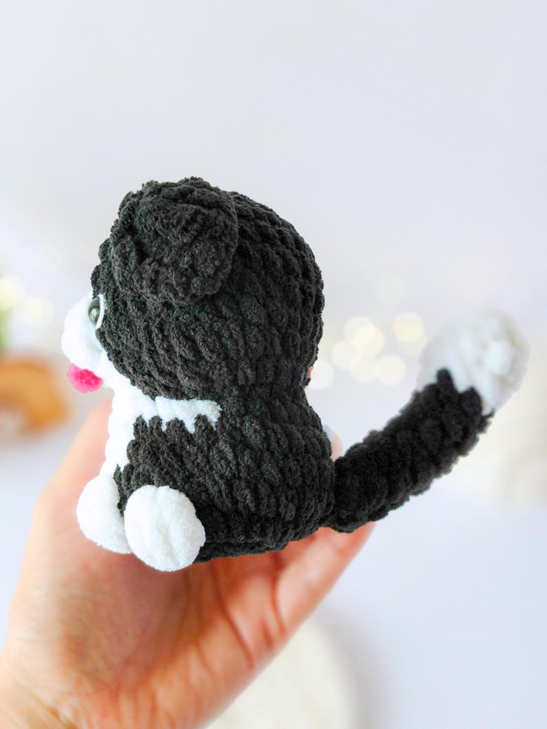 May include: A small, handmade crochet cat plushie featuring a black and white color scheme. The cat has a textured, fuzzy yarn body, a long tail with a white tip, small white paws, and a tiny pink tongue visible on its side profile.