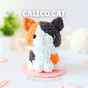 May include: A plush calico cat toy made of chunky yarn with orange, black, and white patches, black safety eyes, and a pink stitched nose. The text CALICO CAT and no-sew crochet pattern are visible above and below the cat on a pink circular base.